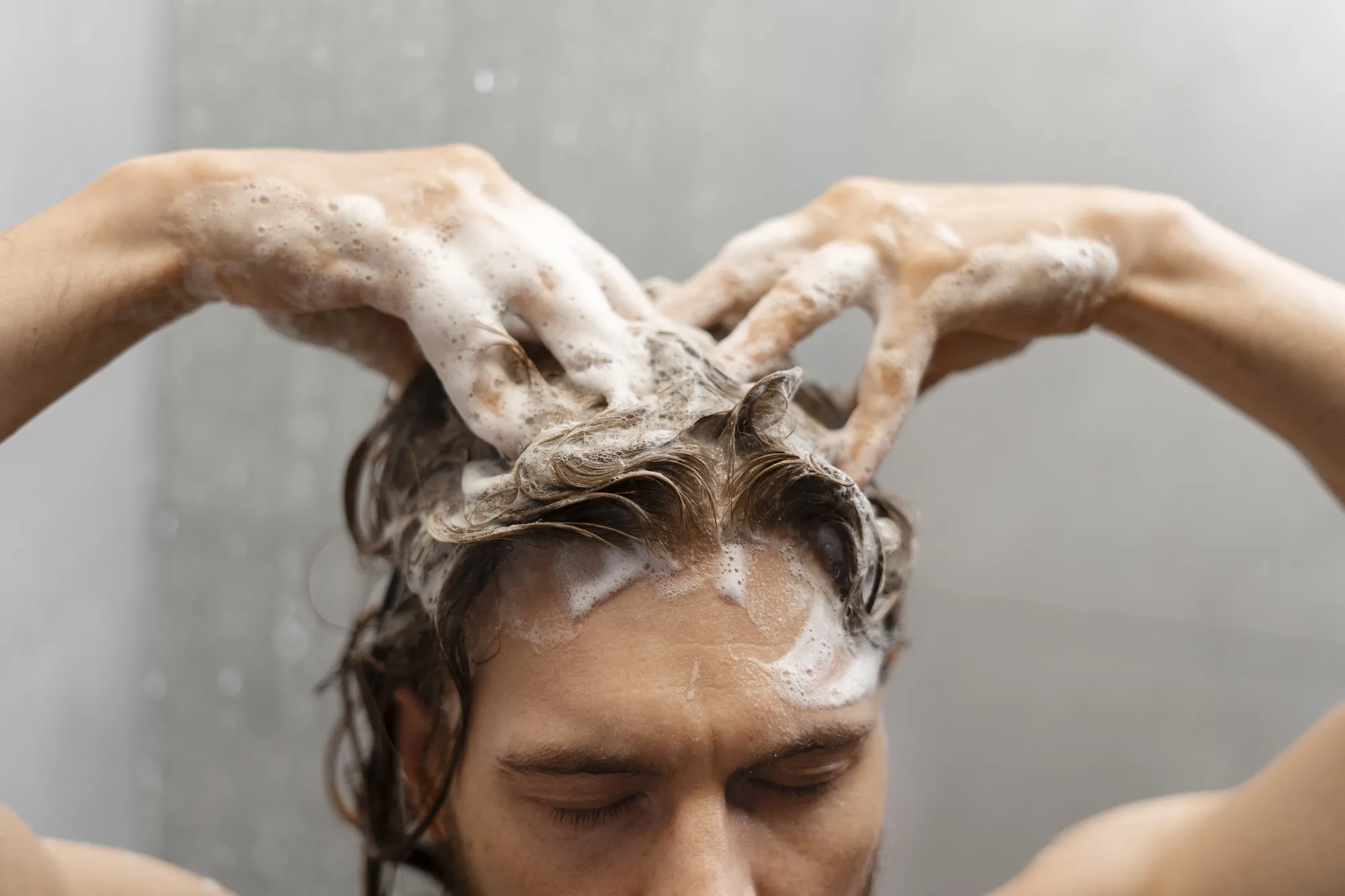 Man onder de douche masseert shampoo met beide handen in zijn haar en hoofdhuid.